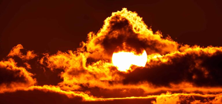 The bright sun, shining orange, with some clouds surrounding it.