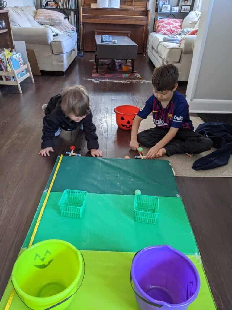 Two kids sitting in front of a green mat, using popsicle sticks to play a game. 